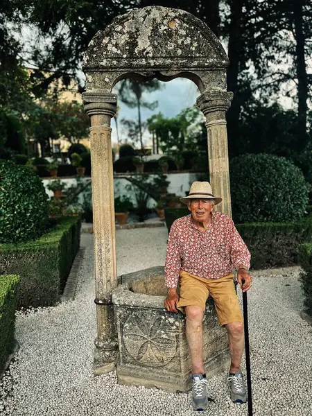 Senior man in red white shirt, straw hat and brown shorts sits on edge of water well in the garden of Casa del Rey Moro in Ronda, Andalucia, Spain.