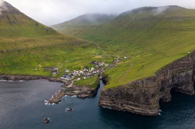 Faroe Adaları 'ndaki yemyeşil dağların arasına kurulmuş görkemli bir köyü yakalayan insansız hava aracı görüntüsü..