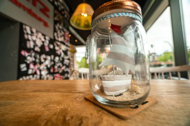 A whimsical miniature sailboat inside a glass jar, beautifully displayed on a rustic wooden table in a vibrant cafe in Hamburg. clipart