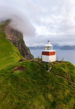 Faroe Adaları 'ndaki manzaralı deniz fenerinin göz kamaştırıcı manzarası canlı yeşil uçurumlar ve sakin bir okyanus zeminiyle çerçevelenmiş..
