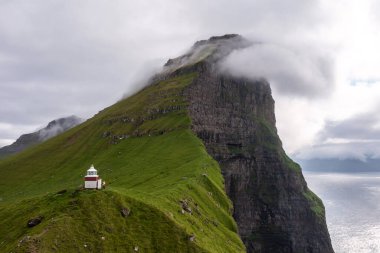Bereketli yeşil uçurumlar ve sisli gökyüzü ile çevrili Faroe Adaları 'ndaki deniz fenerinin çarpıcı görüntüsü seyahat ve doğa temaları için mükemmel..