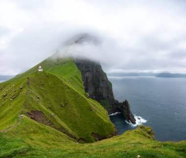 Faroe Adaları 'nın büyüleyici panoramik manzarası yemyeşil uçurumlar ve bulutlu gökyüzünün altında büyüleyici bir deniz feneri sergiliyor..
