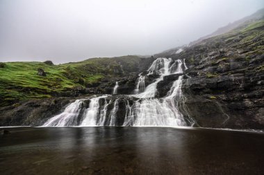 Büyüleyici bir şelale Faroe Adaları 'ndaki kayalık kayalıklardan aşağı süzülüyor, yemyeşil ve sisli dağlarla çevrili..