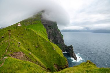 Faroe Adaları 'ndaki bir uçuruma tünemiş bir deniz fenerinin çarpıcı hava manzarası canlı yeşil tepeler ve okyanus ötesini gösteriyor..