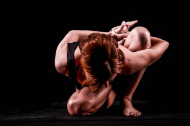 Genç, ince, atletik, siyah tişörtlü ve siyah taytlı kızıl saçlı bir kız siyah bir arka planda, bir fotoğraf stüdyosundaki Asana 'da oturuyor. Yüksek kalite fotoğraf