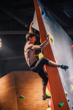 Güzel şişirilmiş vücudu olan genç, atletik bir adam tırmanma salonundaki kayaya tırmanıyor. Yüzünde duygular var. Yüksek kalite fotoğraf