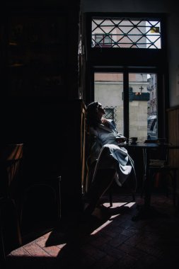 Beyaz elbiseli genç kız güneşin altında pencerenin yanındaki bir kafede kahve içiyor. Siluet. Yüksek kalite fotoğraf