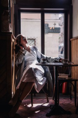 Beyaz elbiseli genç kız güneşin altında pencerenin yanındaki bir kafede kahve içiyor. Siluet. Yüksek kalite fotoğraf
