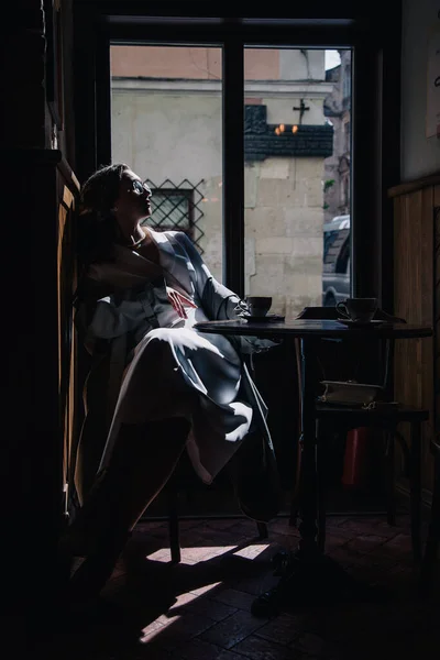 Stock image Young girl in white dress drinking coffee in a coffee shop near the window in the sun. Silhouette. High quality photo