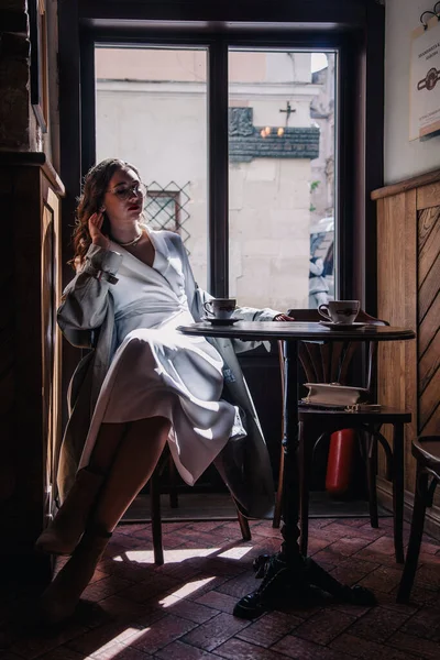 stock image Young girl in white dress drinking coffee in a coffee shop near the window in the sun. Silhouette. High quality photo