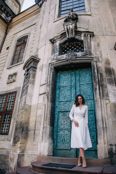 stock image A young, beautiful girl in a white dress and a gray cloak poses near the ancient gray doors of the temple. High quality photo