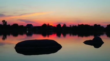 İnanılmaz günbatımı renkleri, göl, sudaki taşlar, küçük dalgalar, ağaç siluetleri.
