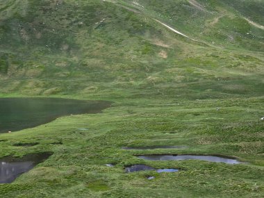 La Thuile, Val D 'Aosta, Temmuz 02 2025: Alplere panoramik manzara, küçük doğal bir göle yansıyan güzel yeşil manzara. Bulutlu güzel mavi gökyüzü. Biraz karlı dağlar. La Thuile köyünün havadan görünüşü.