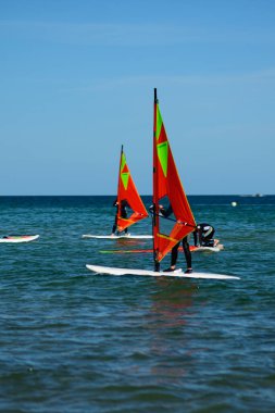 Deniz ve insanlar rüzgar sörfü yapıyor. Parlak kırmızı ve turuncu sarı yelkenli gemilerin üzerinde duruyorlar. Su sakin ve gökyüzü parlak mavi, küçük bir bulutla. Aksiyon açık ve güneşli bir günde gerçekleşir.