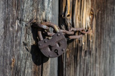 A close-up of an old rusty metal padlock on a weathered wooden door. Sunlight casts shadows, highlighting rust, texture, and chain details, creating a nostalgic, abandoned, and atmospheric scene.