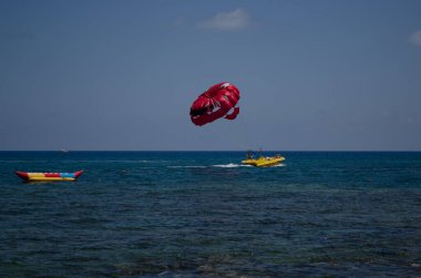 Mavi denizin üzerinde uçan parlak kırmızı paraşüt, güneşli bir yaz gününde sarı bir sürat teknesi tarafından çekiliyor, su sporları macera konsepti.