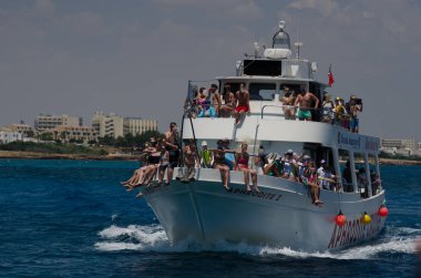 Güneşli bir yaz gününde gezen turistlerle dolu bir tekne, el sallayan ve fotoğraf çeken insanlar, arka planda temiz mavi su ve sahil manzarası. Kıbrıs 11.06.2014