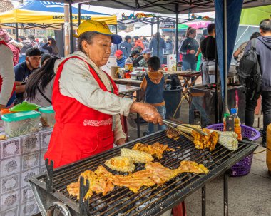 Cuenca, Ekvador - 7 Temmuz 2024 Cuenca kentinde et festivali. Bir kadın ızgarada tavuk bifteği ve mısır koçanı pişiriyor. Sokak yemeği.