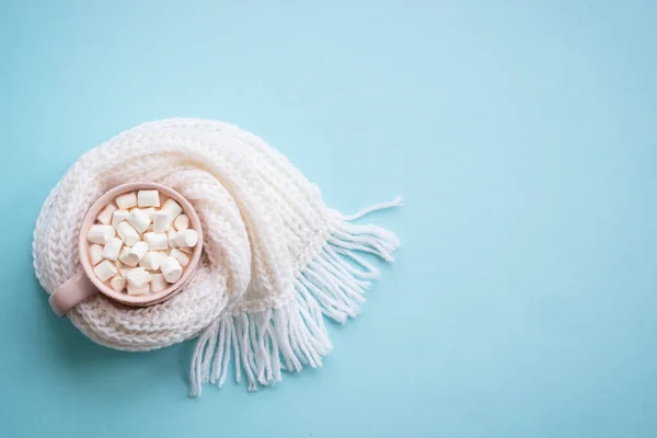 Hot chocolate with marshmallows and knitting scarf on blue. Cozy winter concept, flat lay image.