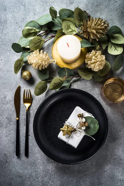 stock image Christmas table setting with black plate, candles, eucalyptus leaves and golden decorations. Top view at stone table.