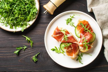 Krem peynirli sandviç, prosciutto, salatalık ve roka beyaz tabakta ahşap arka planda..