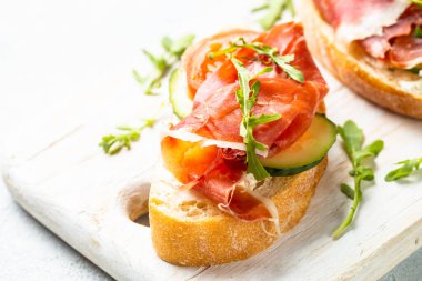 Beyaz arka planda kesme tahtasının üzerinde prosciutto ve roka ile sandviç açın. Resmi kapat.