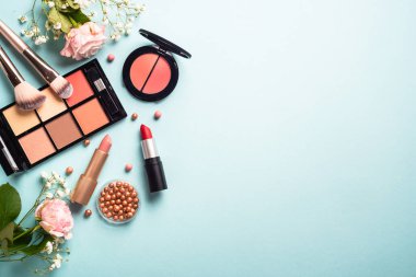 Cosmetic products on blue background. Cream, powder, shadow, brushes with green leaves and flowers. Flat lay image with copy space.