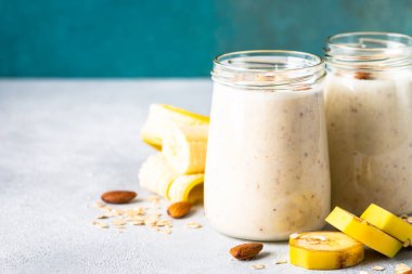 Almond banana smoothie with oat flakes and milk in glass jars at white table.