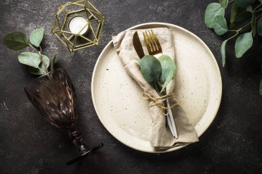 White craft plate, cutlery, wine glass and eucaliptus at black stone table. Top view image.