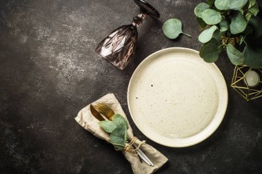 Table setting at black table. White craft plate, cutlery, wine glass and eucaliptus. Top view image.