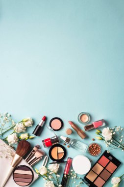 Cosmetic products on blue background. Cream, powder, shadow, brushes with green leaves and flowers. Flat lay image with copy space.