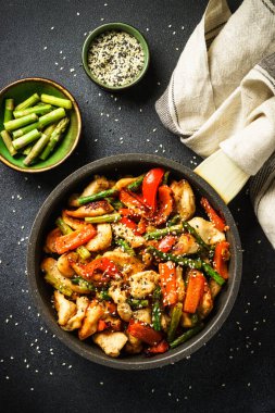 Chicken stir fry with vegetables and sesame in the skillet at black stone background. Top view, vertical.