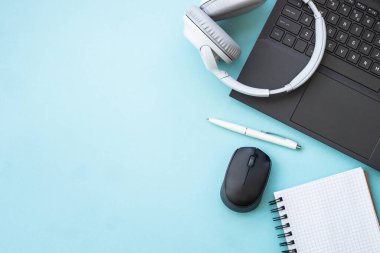 Office desk with laptop, headphones, notepad and pen. Flat lay image on blue with copy space.