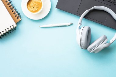 Office workspace with laptop, headphones, notepad and coffee cup. Flat lay image on blue with copy space.