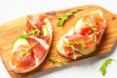 Open sandwiches with cream cheese, prosciutto, cucumber and arugula. Top view on white.