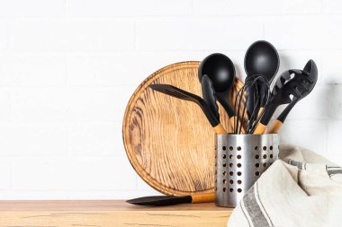 Kitchen utensils, cooking tools in container with wooden cutting board near white wall.