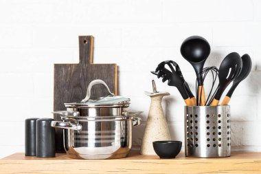 Kitchen table with kitchen utensils, cooking pots, oil bottle with wooden cutting board, white modern interior.