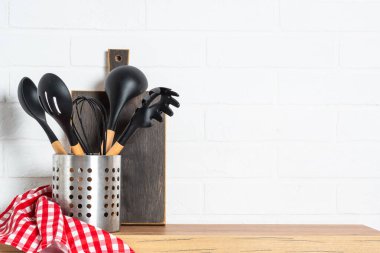 Kitchen utensils with wooden cutting board and towel near white wall.