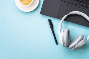 Office desk with laptop, headphones, notepad and pen. Flat lay image on blue with copy space.