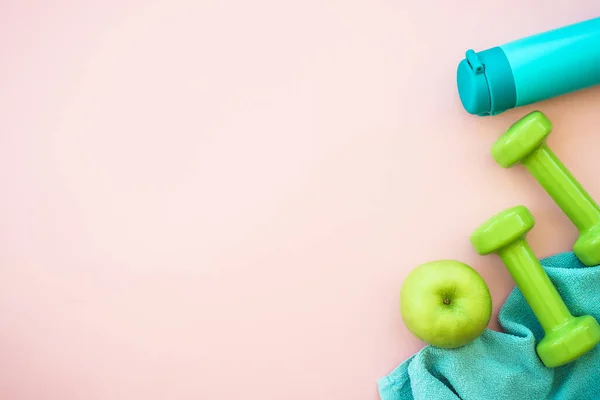 Fitness background on pink. Dumbbells, towel and bottle of water with green apple. Flat lay image with copy space.