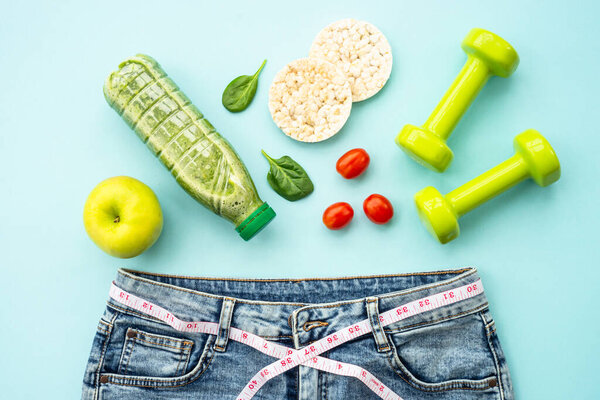 Losing weight background. Dumbells, healthy food and jeans with measuring tape. Flat lay on blue.