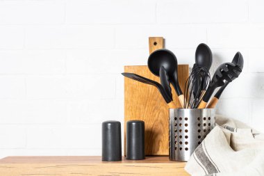 Kitchen utensils, cooking tools in container with wooden cutting board near white wall.