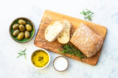 Tahtada ciabatta ekmeği, zeytin yağı, zeytin ve bitkilerle birlikte. Beyaz masadaki üst görünüm.