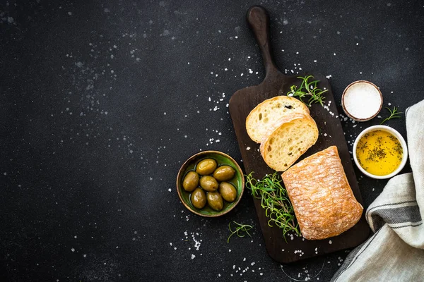 stock image Ciabatta bread on wooden board with olive oil, olives and herbs on black. Top view with copy space.