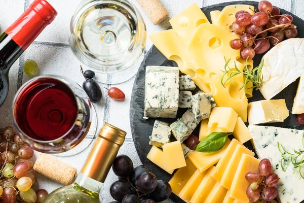 Cheese platter with craft cheese assortment and wine glasses at white tile background. Top view.