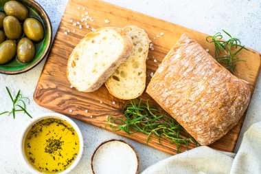 Tahtada ciabatta ekmeği, zeytin yağı, zeytin ve bitkilerle birlikte. Beyaz masadaki üst görünüm.