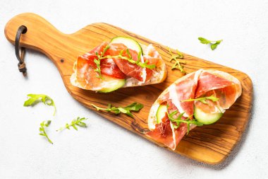 Open sandwiches with cream cheese, prosciutto, cucumber and arugula. Top view on white.
