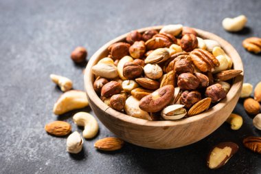 Koyu taş masadaki ahşap kasede fındık çeşitleri. Kaju, fındık, badem, Brezilya fındığı ve ceviz. Kapat..