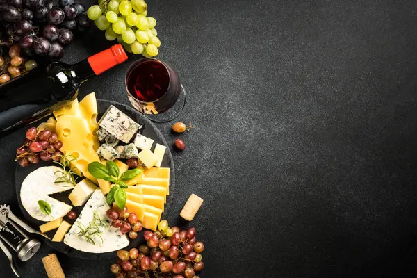 Cheese platter with craft cheese assortment on slate board and red wine at black background. Top view with copy space.