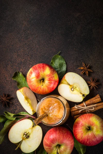 Apple jam in the glass jar and fresh apples, leaves and spices on dark stone background. Top view.